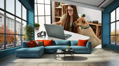 Young hispanic woman using laptop sitting on table with dog at home Wall mural