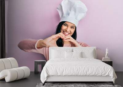 Young hispanic woman wearing baker uniform and cook hat smiling in love showing heart symbol and shape with hands. romantic concept. Wall mural