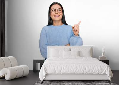 Young hispanic woman wearing casual clothes and glasses with a big smile on face, pointing with hand and finger to the side looking at the camera. Wall mural