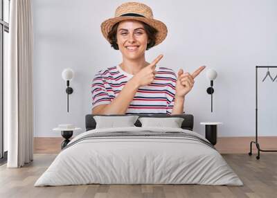 Young hispanic woman wearing casual clothes and hat sitting on the table smiling and looking at the camera pointing with two hands and fingers to the side. Wall mural