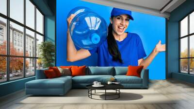 Young hispanic woman wearing delivery uniform holding water carafe celebrating achievement with happy smile and winner expression with raised hand Wall mural