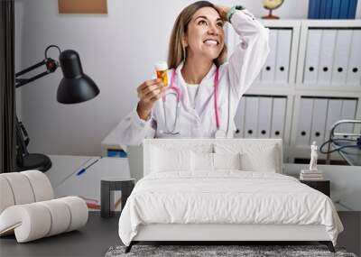 Young hispanic woman wearing doctor uniform holding pills at the clinic smiling confident touching hair with hand up gesture, posing attractive and fashionable Wall mural