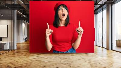 Young hispanic woman wearing french look with beret amazed and surprised looking up and pointing with fingers and raised arms. Wall mural