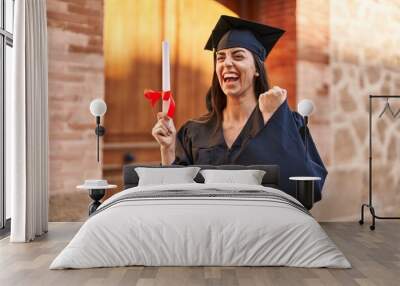 Young hispanic woman wearing graduated uniform holding diploma at university Wall mural