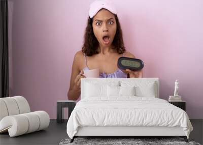 Young hispanic woman wearing nightgown holding alarm clock in shock face, looking skeptical and sarcastic, surprised with open mouth Wall mural