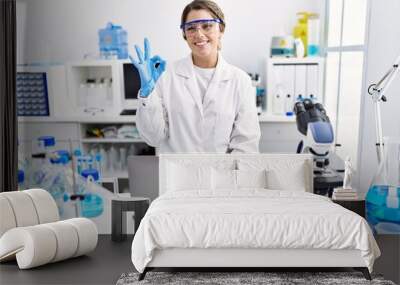 Young hispanic woman wearing scientist uniform working at laboratory doing ok sign with fingers, smiling friendly gesturing excellent symbol Wall mural