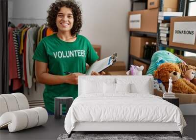 Young hispanic woman wearing volunteer t shirt at donations stand holding checklist looking positive and happy standing and smiling with a confident smile showing teeth Wall mural