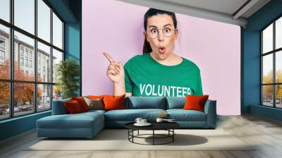 Young hispanic woman wearing volunteer t shirt surprised pointing with finger to the side, open mouth amazed expression. Wall mural