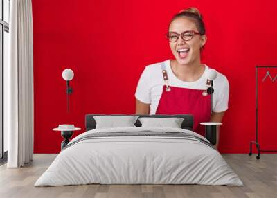 Young hispanic woman wearing waitress apron over red background winking looking at the camera with sexy expression, cheerful and happy face. Wall mural