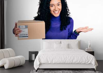 Young hispanic woman with curly hair holding delivery package celebrating achievement with happy smile and winner expression with raised hand Wall mural