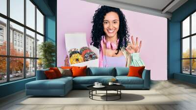 Young hispanic woman with curly hair holding tasty colorful doughnuts box doing ok sign with fingers, smiling friendly gesturing excellent symbol Wall mural