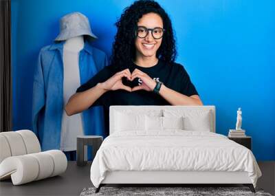 Young hispanic woman with curly hair wearing staff t shirt smiling happy doing ok sign with hand on eye looking through fingers Wall mural