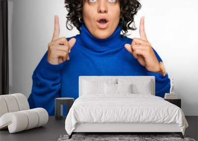 Young hispanic woman with curly hair wearing turtleneck sweater amazed and surprised looking up and pointing with fingers and raised arms. Wall mural