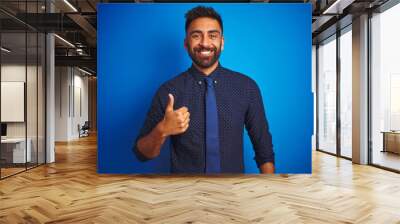 Young indian businessman wearing elegant shirt and tie standing over isolated blue background doing happy thumbs up gesture with hand. Approving expression looking at the camera showing success. Wall mural