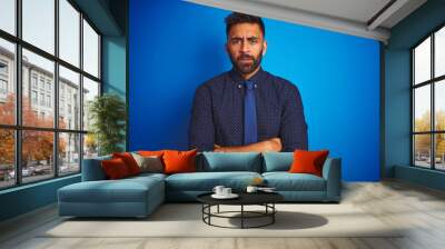 Young indian businessman wearing elegant shirt and tie standing over isolated blue background skeptic and nervous, disapproving expression on face with crossed arms. Negative person. Wall mural