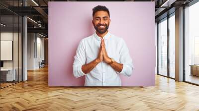 Young indian businessman wearing elegant shirt standing over isolated pink background praying with hands together asking for forgiveness smiling confident. Wall mural