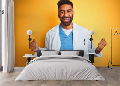 Young indian doctor man standing over isolated yellow background celebrating surprised and amazed for success with arms raised and open eyes. Winner concept. Wall mural