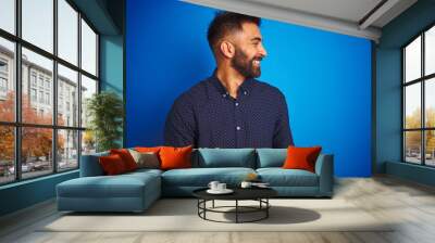 Young indian elegant man wearing shirt standing over isolated blue background looking away to side with smile on face, natural expression. Laughing confident. Wall mural