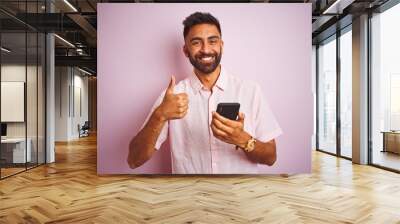 Young indian man using smartphone standing over isolated pink background happy with big smile doing ok sign, thumb up with fingers, excellent sign Wall mural