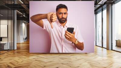 Young indian man using smartphone standing over isolated pink background with angry face, negative sign showing dislike with thumbs down, rejection concept Wall mural