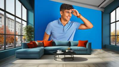 Young indian man wearing casual polo standing over isolated blue background very happy and smiling looking far away with hand over head. Searching concept. Wall mural