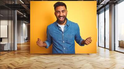 Young indian man wearing denim shirt standing over isolated yellow background celebrating surprised and amazed for success with arms raised and open eyes. Winner concept. Wall mural