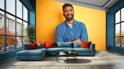 Young indian man wearing denim shirt standing over isolated yellow background happy face smiling with crossed arms looking at the camera. Positive person. Wall mural