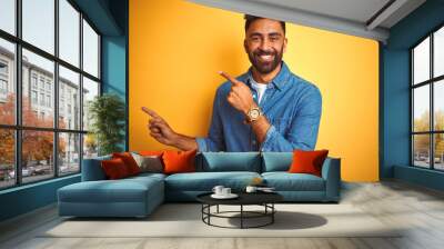 Young indian man wearing denim shirt standing over isolated yellow background smiling and looking at the camera pointing with two hands and fingers to the side. Wall mural