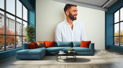 Young indian man wearing elegant shirt standing over isolated white background looking away to side with smile on face, natural expression. Laughing confident. Wall mural