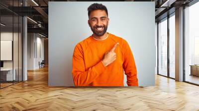 Young indian man wearing orange sweater over isolated white background cheerful with a smile of face pointing with hand and finger up to the side with happy and natural expression on face Wall mural