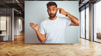 Young indian man wearing t-shirt standing over isolated white background confused and annoyed with open palm showing copy space and pointing finger to forehead. Think about it. Wall mural