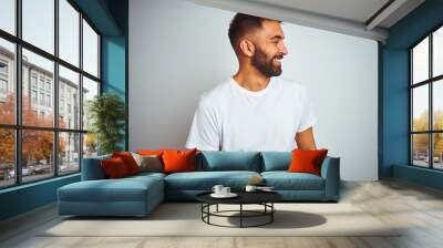 Young indian man wearing t-shirt standing over isolated white background looking away to side with smile on face, natural expression. Laughing confident. Wall mural