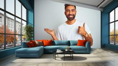 Young indian man wearing t-shirt standing over isolated white background Showing palm hand and doing ok gesture with thumbs up, smiling happy and cheerful Wall mural