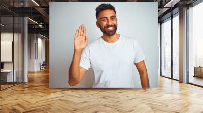 Young indian man wearing t-shirt standing over isolated white background Waiving saying hello happy and smiling, friendly welcome gesture Wall mural