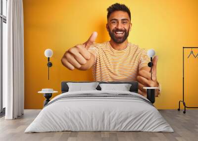 Young indian man wearing t-shirt standing over isolated yellow background approving doing positive gesture with hand, thumbs up smiling and happy for success. Winner gesture. Wall mural