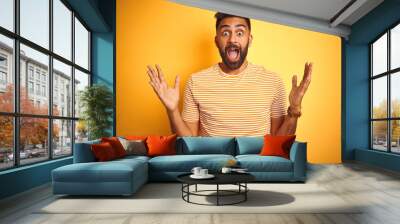 Young indian man wearing t-shirt standing over isolated yellow background celebrating crazy and amazed for success with arms raised and open eyes screaming excited. Winner concept Wall mural