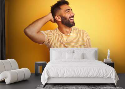 Young indian man wearing t-shirt standing over isolated yellow background smiling confident touching hair with hand up gesture, posing attractive and fashionable Wall mural