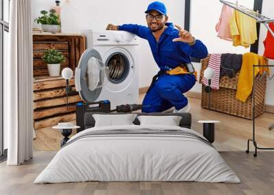 Young indian technician working on washing machine smiling and confident gesturing with hand doing small size sign with fingers looking and the camera. measure concept. Wall mural