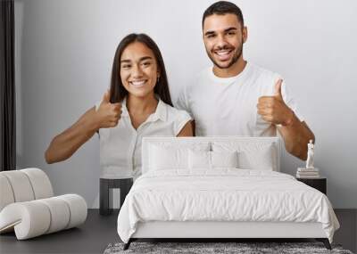 Young interracial couple standing together in love over isolated background doing happy thumbs up gesture with hand. approving expression looking at the camera showing success. Wall mural