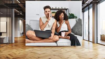 Young interracial couple using laptop at home sitting on the sofa hand on mouth telling secret rumor, whispering malicious talk conversation Wall mural