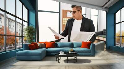 Young latin businessman with serious expression reading paperwork standing at the office. Wall mural