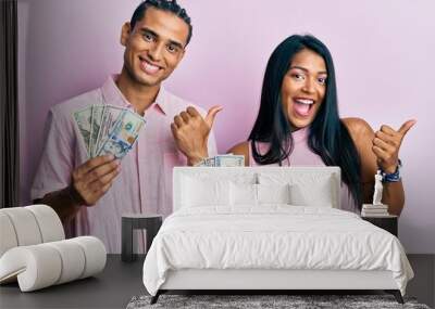 Young latin couple holding dollars pointing thumb up to the side smiling happy with open mouth Wall mural