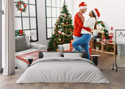 Young latin couple hugging and standing by christmas tree at home. Wall mural