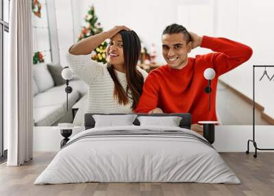 Young latin couple sitting on the table by christmas tree smiling confident touching hair with hand up gesture, posing attractive and fashionable Wall mural