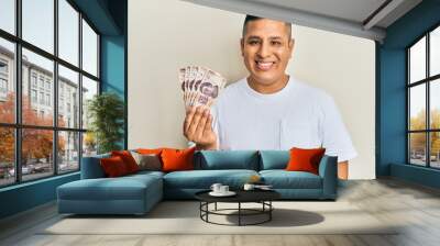 Young latin man holding 500 mexican pesos banknotes looking positive and happy standing and smiling with a confident smile showing teeth Wall mural