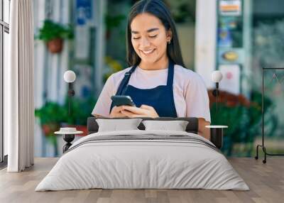 Young latin shopkeeper girl smiling happy using smartphone at florist. Wall mural