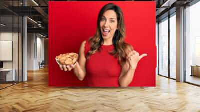 Young latin woman holding bowl with salty biscuits pointing thumb up to the side smiling happy with open mouth Wall mural