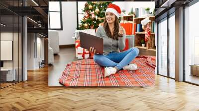 Young latin woman using laptop sitting by christmas tree doing money gesture with hands, asking for salary payment, millionaire business Wall mural