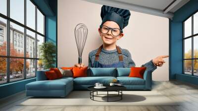 Young little caucasian cook kid wearing chef uniform and hat using manual whisk very happy pointing with hand and finger to the side Wall mural