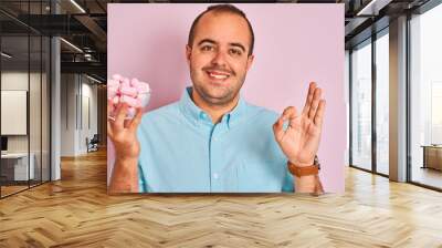 Young man holding bowl with marshmallows standing over isolated pink background doing ok sign with fingers, excellent symbol Wall mural